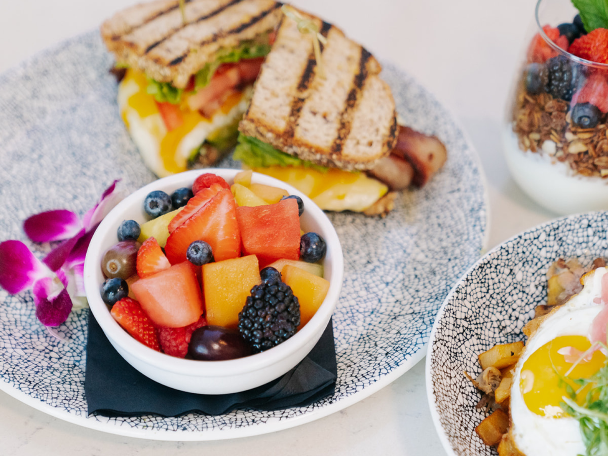 The image shows a sandwich, a fruit bowl, sunny-side-up eggs with greens, and a yogurt parfait topped with berries and granola.