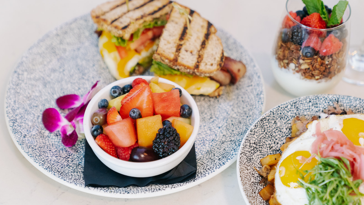 The image shows a sandwich, a fruit bowl, sunny-side-up eggs with greens, and a yogurt parfait topped with berries and granola.