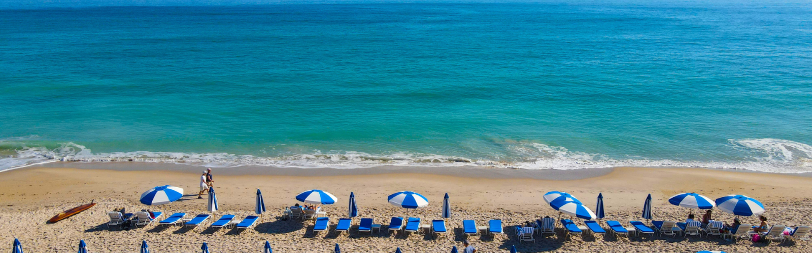 A serene beach scene with clear blue water, rows of blue and white umbrellas, and beach chairs on the sand, with a few people relaxing.