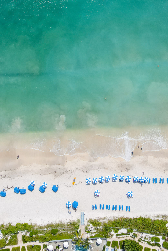 This aerial view shows a beach with blue umbrellas, lounge chairs, and a few people, bordered by lush greenery, and the ocean with gentle waves.