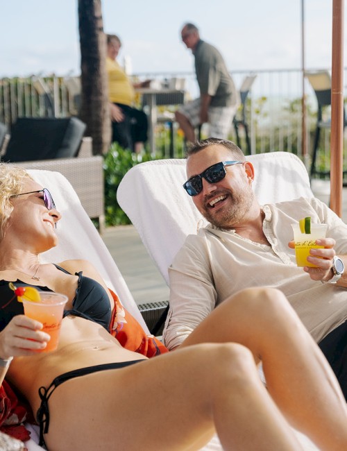 Two friends enjoy drinks and laughter while relaxing on sun loungers at a poolside or beach club.