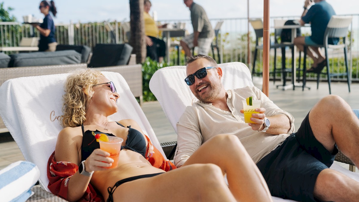 Two people are lounging on chairs by a pool, enjoying drinks and smiling at each other with more people in the background.