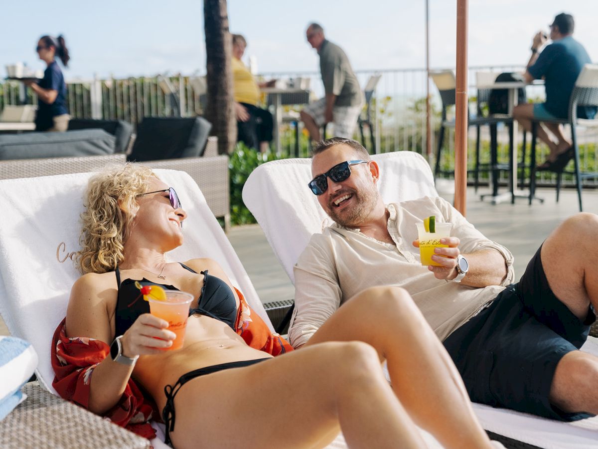 Two people are lounging on chairs by a pool, enjoying drinks and smiling at each other with more people in the background.