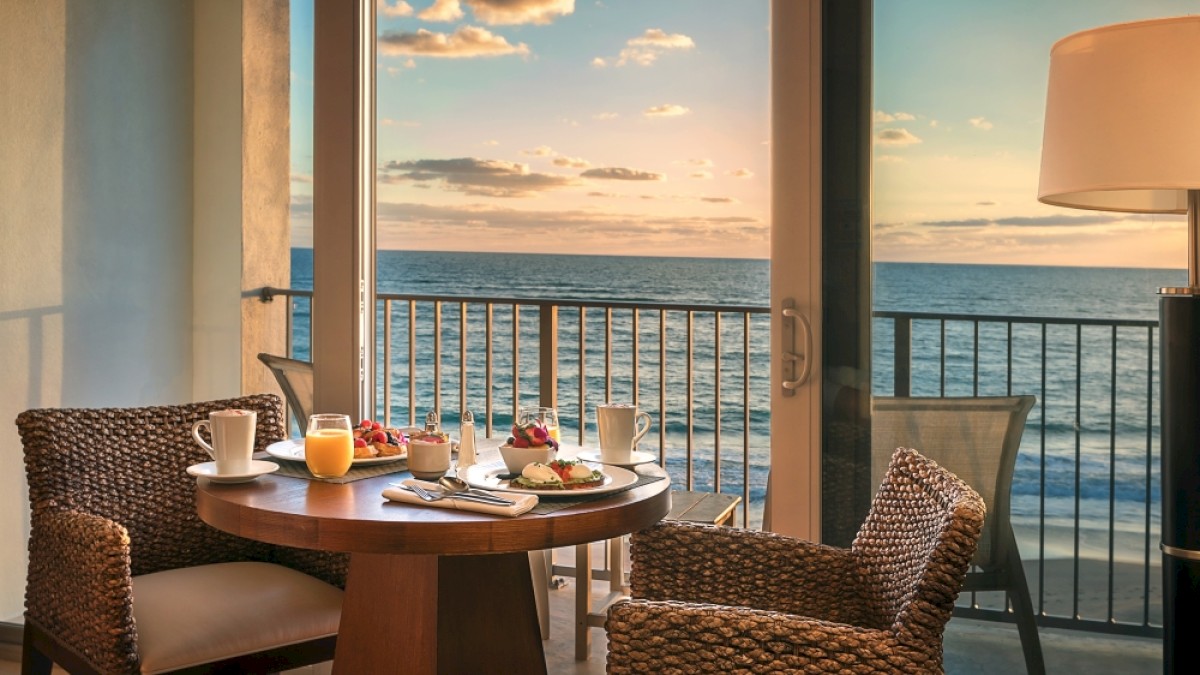 A cozy seaside breakfast setup with coffee, juice, and pastries on a round table overlooking the ocean at sunrise.