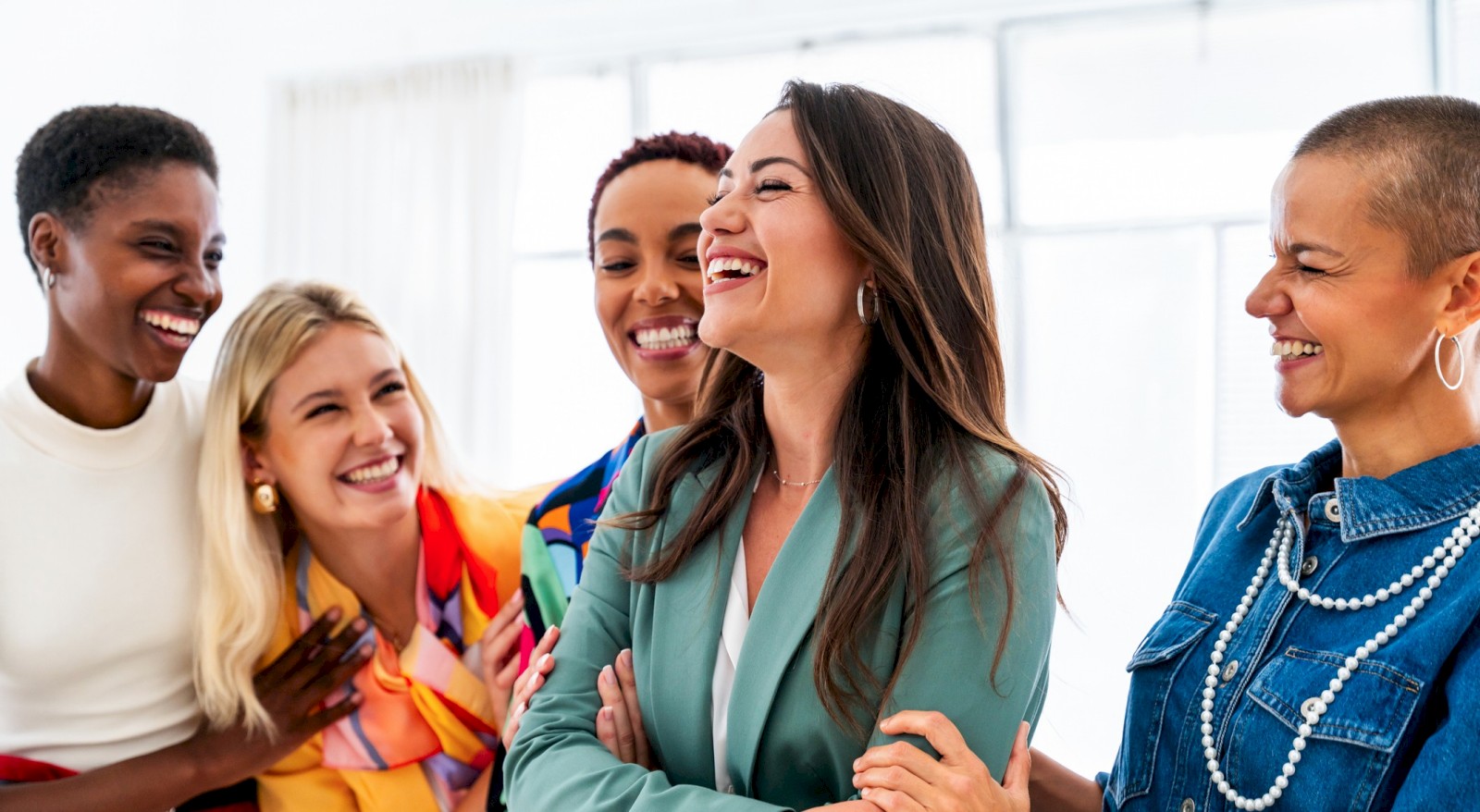 A diverse group of women sharing a joyful, embracing moment indoors with big smiles and laughter.