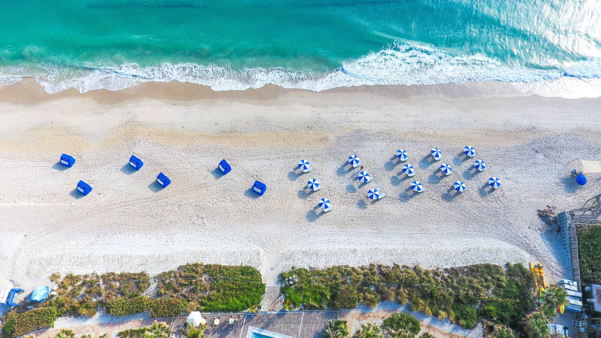 An aerial view of a beach with turquoise water, white sand, blue umbrellas, and beach chairs, surrounded by greenery and buildings.