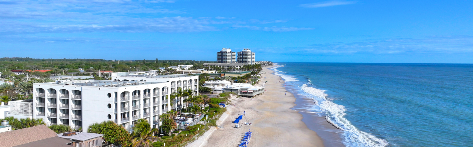Aerial view of a beach with resorts, a sandy shoreline, beach chairs, and the ocean under a clear blue sky.