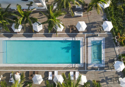 The image shows an aerial view of a swimming pool area with lounge chairs, umbrellas, and palm trees surrounding the pool on a wooden deck.