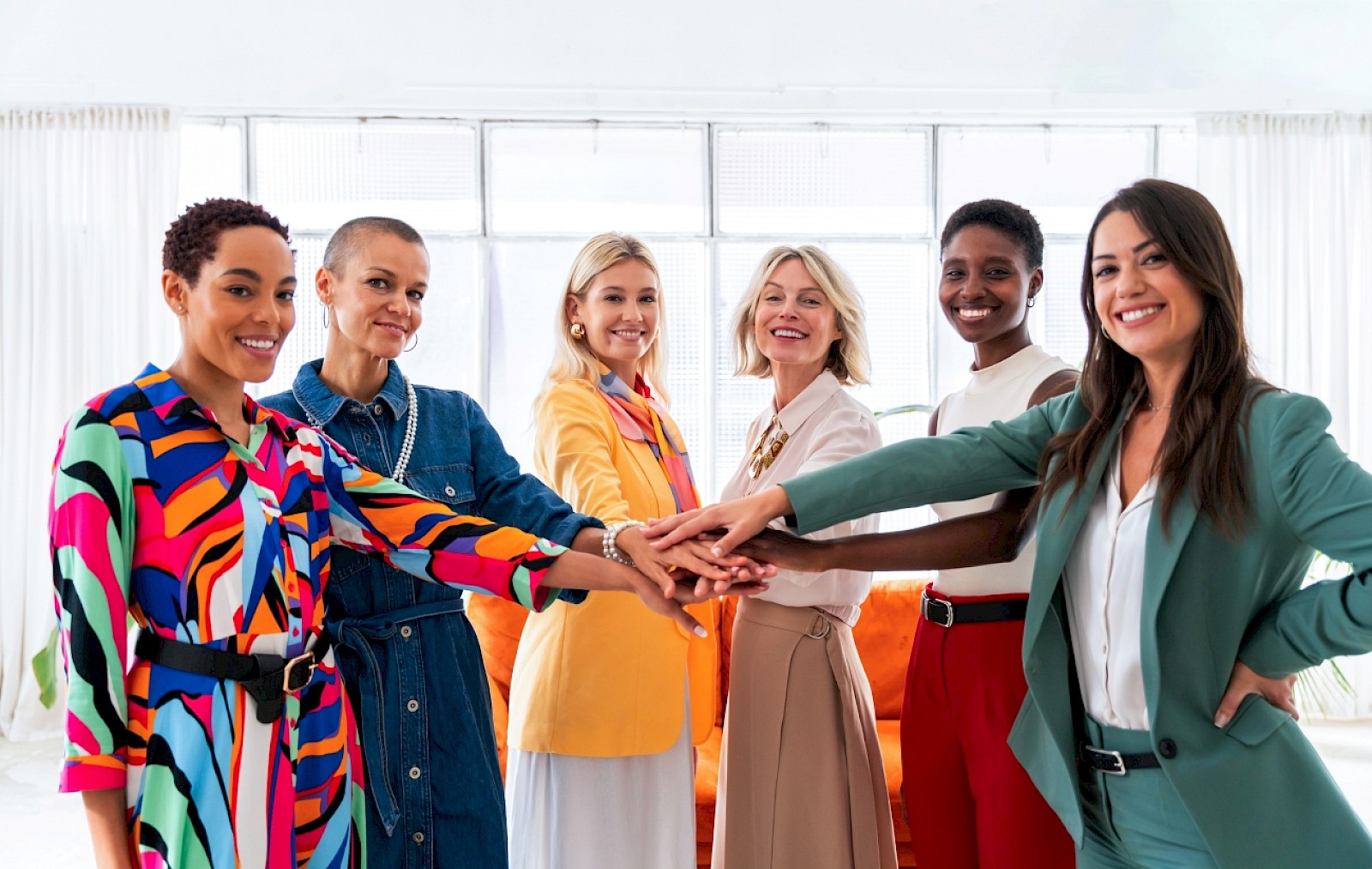 Seven diverse women in bright, colorful outfits are smiling and putting their hands together in a show of teamwork and unity.