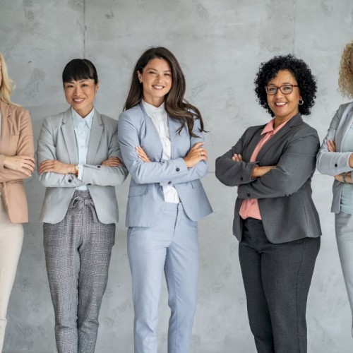 A diverse group of five confident women in business attire standing together against a gray wall.