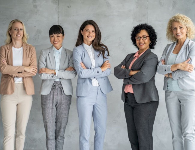 A diverse group of five confident women in business attire standing together against a gray wall.
