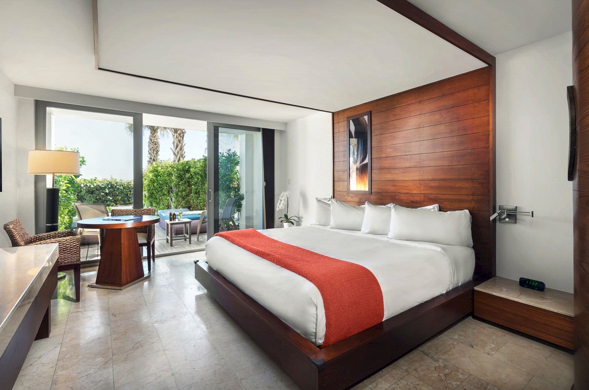 A modern hotel room with a large bed, red throw, artwork above the headboard, seating area by the window, and patio with outdoor furniture.