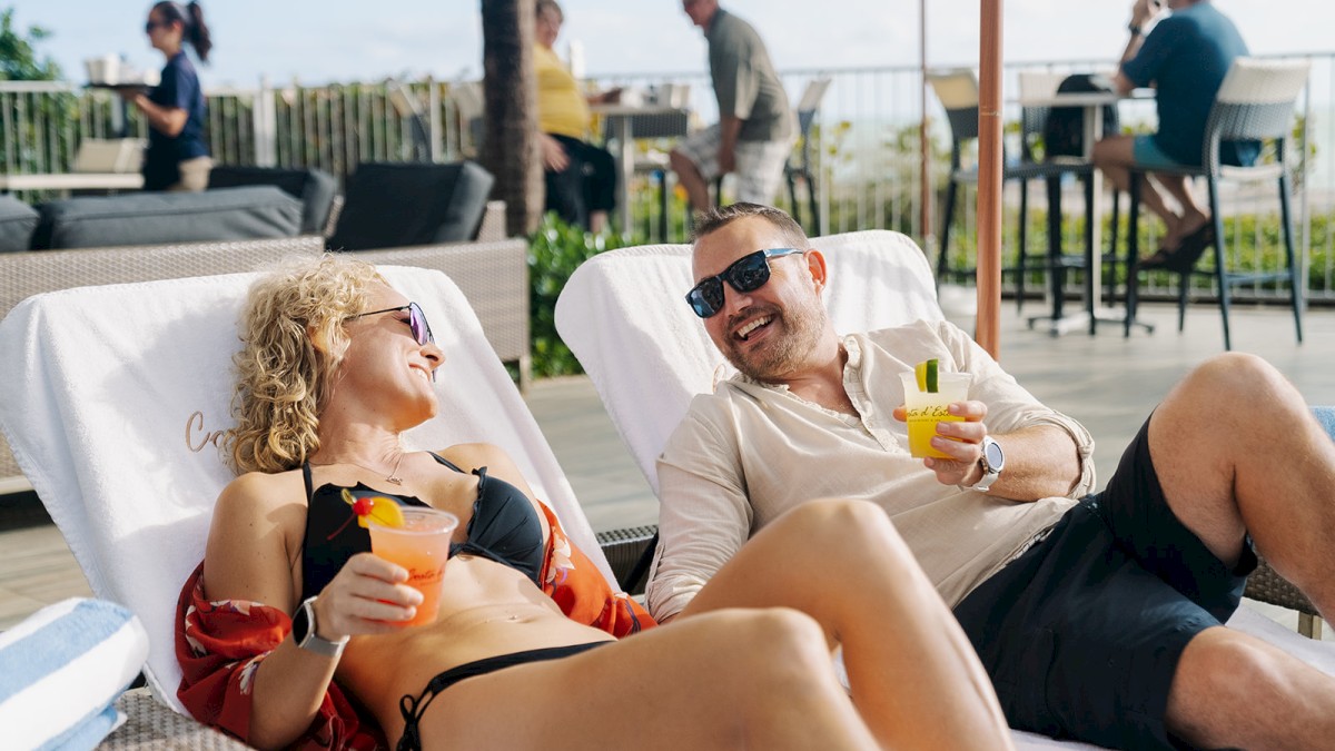 Two people relax on lounge chairs, smiling and holding cocktails. Other individuals are in the background, enjoying what seems to be a poolside area.
