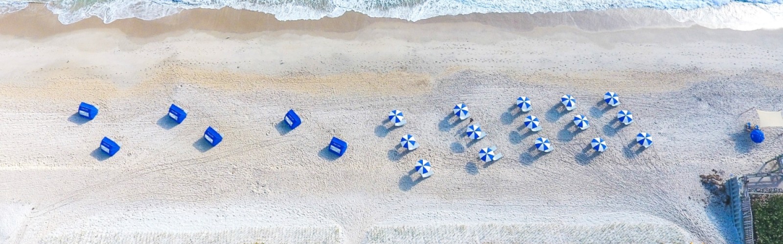 This image shows an aerial view of a beach with turquoise water, white sand, blue umbrellas, and lounge chairs, bordered by vegetation and a swimming pool.