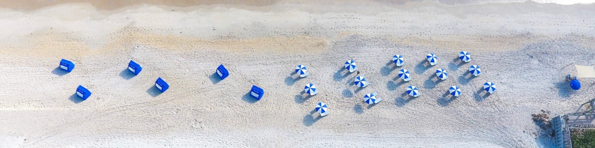 This image shows an aerial view of a beach with turquoise water, white sand, blue umbrellas, and lounge chairs, bordered by vegetation and a swimming pool.