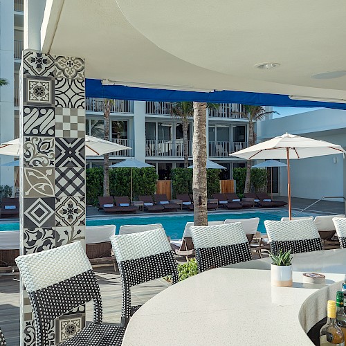 A view of a hotel pool area with lounge chairs, umbrellas, palm trees, and a bar with bottles.