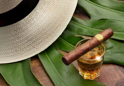 A straw hat, a glass of whiskey with a cigar on top, and large tropical leaves are arranged on a wooden surface.