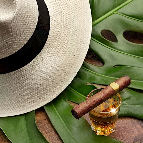 A straw hat, a glass of whiskey with a cigar on top, and large tropical leaves are arranged on a wooden surface.