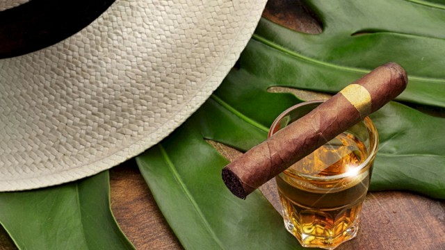 A straw hat, a glass of whiskey with a cigar on top, and large tropical leaves are arranged on a wooden surface.