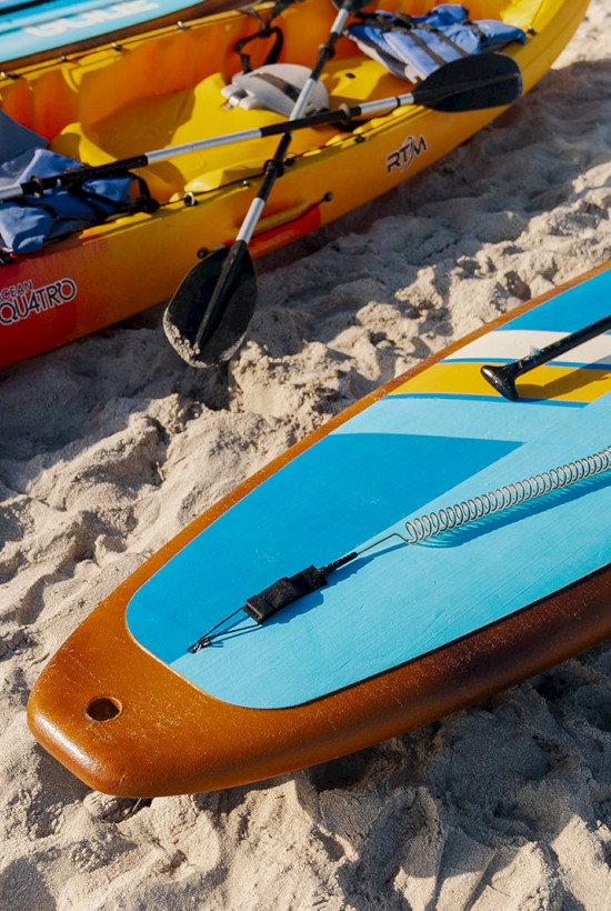 Two kayaks and a paddleboard are on the sandy beach, with paddles and life jackets visible.