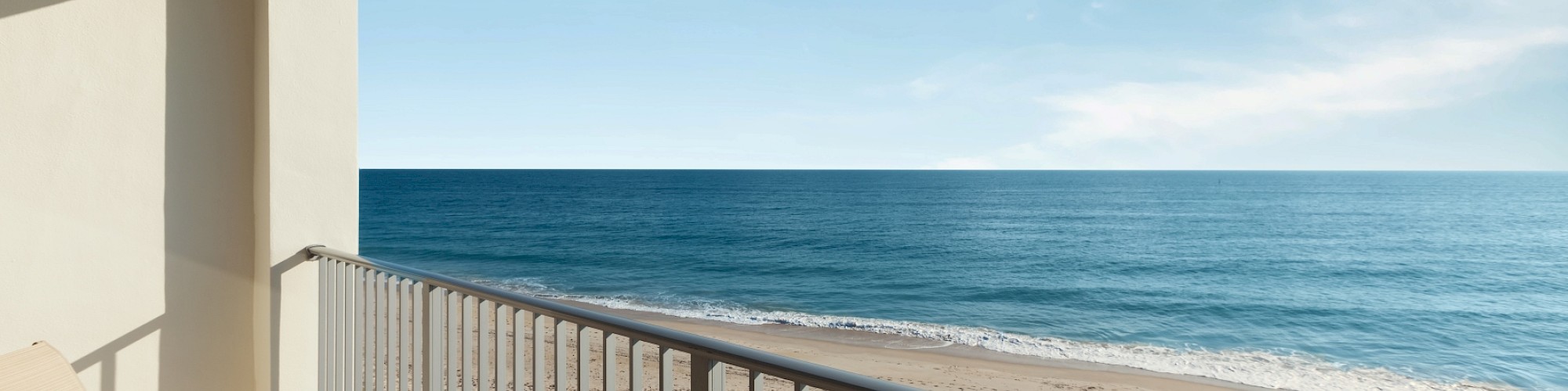 A balcony with chairs overlooks a sandy beach and the calm ocean, under a clear blue sky with a few clouds.