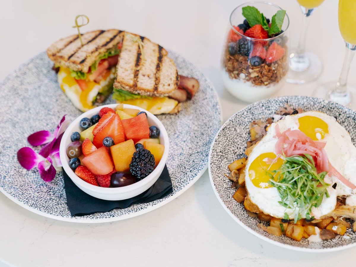 The image shows a well-presented brunch with a grilled sandwich, a bowl of fresh fruit, a dish with eggs and veggies, and parfait and drinks.