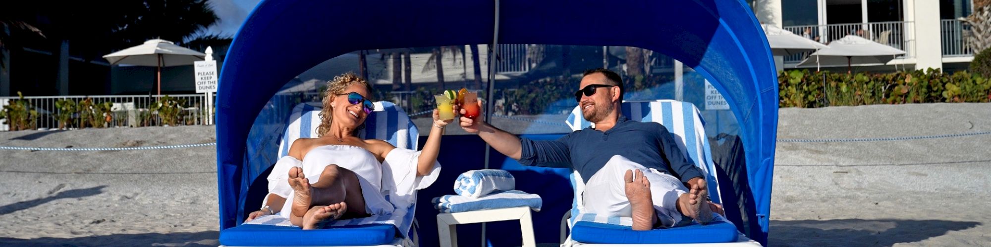Two people are relaxing on beach chairs under a canopy, toasting drinks, with a beach and building in the background.