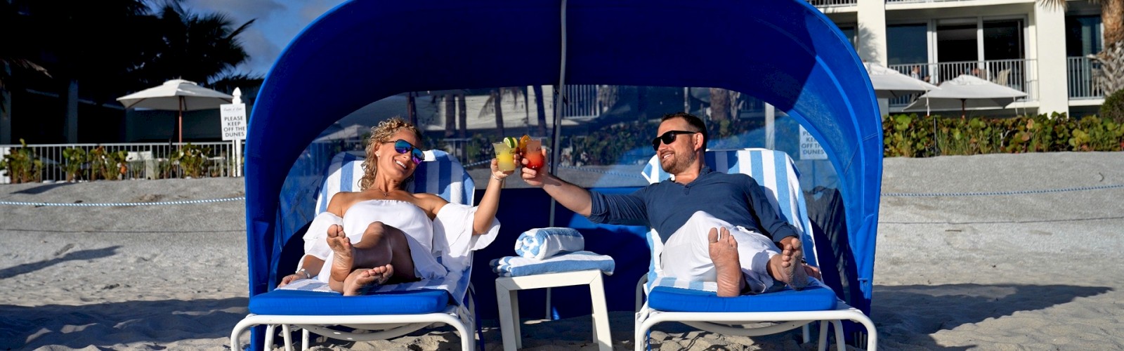 Two people are relaxing on beach chairs under a canopy, toasting drinks, with a beach and building in the background.