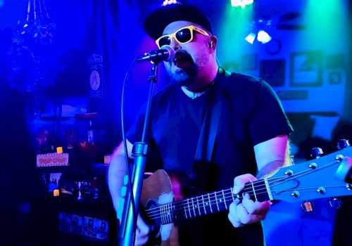 A man wearing sunglasses, a hat, and a black shirt, playing an acoustic guitar and singing into a microphone in a colorful, dimly lit room.