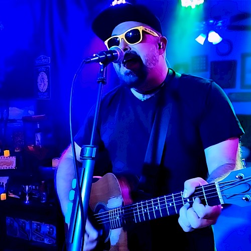 A man wearing sunglasses, a hat, and a black shirt, playing an acoustic guitar and singing into a microphone in a colorful, dimly lit room.