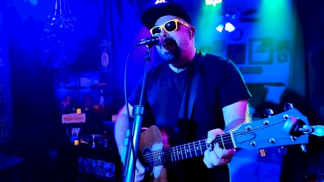 A man wearing sunglasses, a hat, and a black shirt, playing an acoustic guitar and singing into a microphone in a colorful, dimly lit room.
