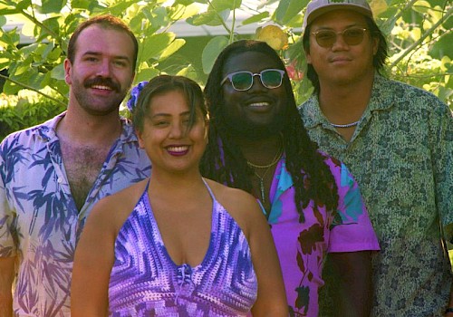 A group of four friends standing outdoors among lush green plants, smiling and enjoying a sunny day.
