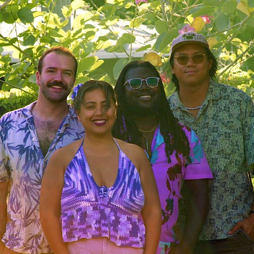 A group of four friends standing outdoors among lush green plants, smiling and enjoying a sunny day.
