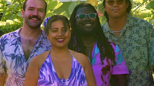 A group of four friends standing outdoors among lush green plants, smiling and enjoying a sunny day.