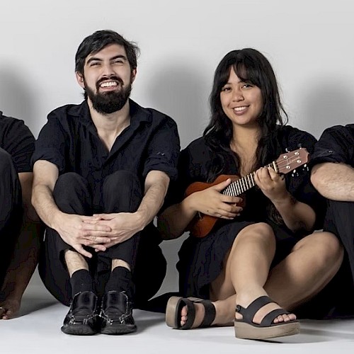 Four happy friends, two men and two women, sit on the floor against a plain background, with one woman playing a ukulele.