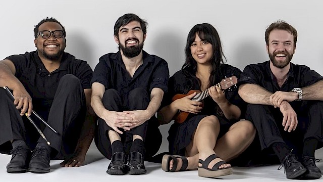 Four happy friends, two men and two women, sit on the floor against a plain background, with one woman playing a ukulele.