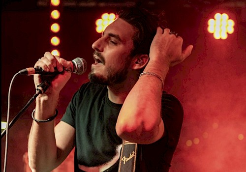 A man with a guitar and a microphone performs passionately on stage under red lights.