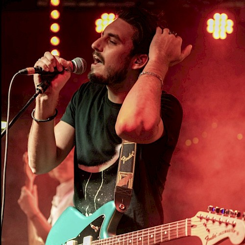 A man with a guitar and a microphone performs passionately on stage under red lights.
