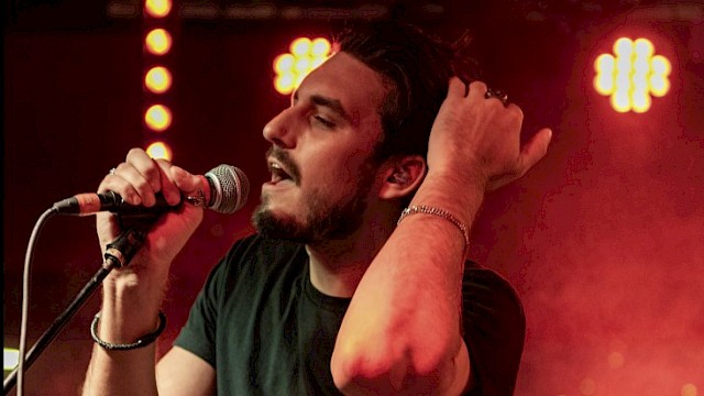 A man with a guitar and a microphone performs passionately on stage under red lights.