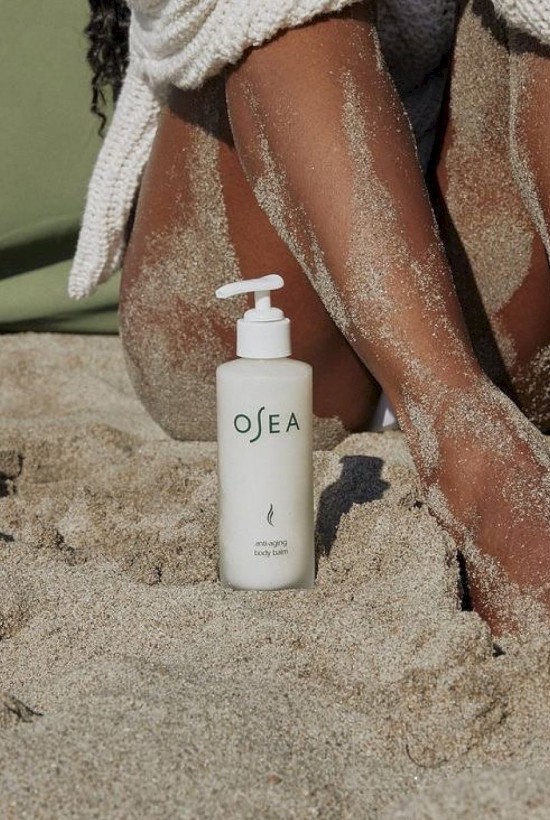A white OSEA body balm bottle sits in sandy beach, with a person's legs covered in sand, beside them.