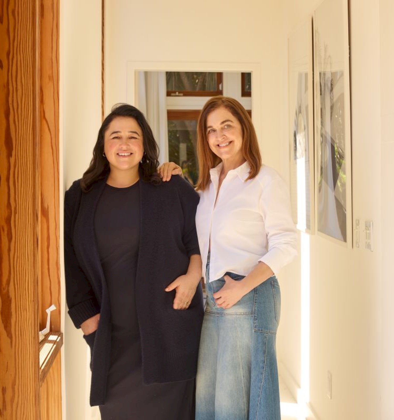 Two women, one in black and one in white, stand together smiling in a bright, cozy home interior with books and plants.