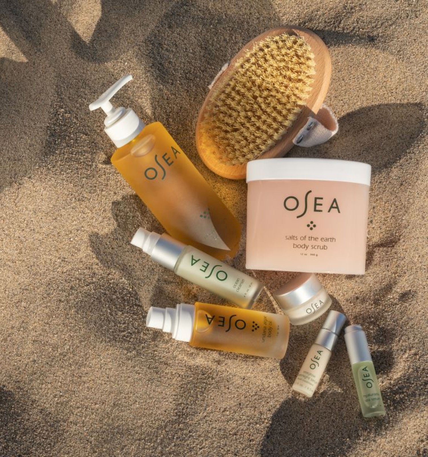 Beach-themed skincare products with a brush, scrub, and serums are laid on sandy ground.