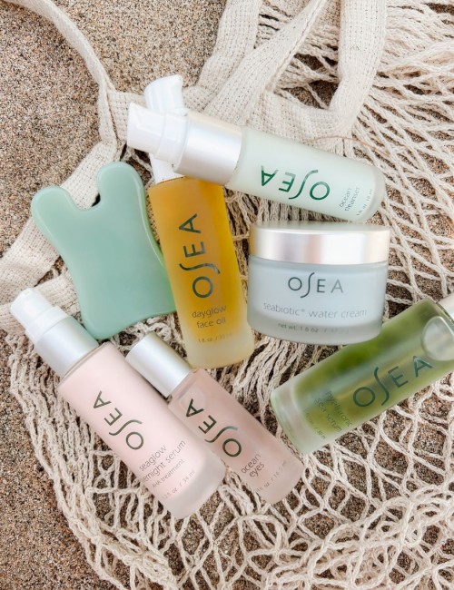 Ocean-themed skincare products are displayed on sand with a net background, including creams, serums, and face oils from the brand OSEA.
