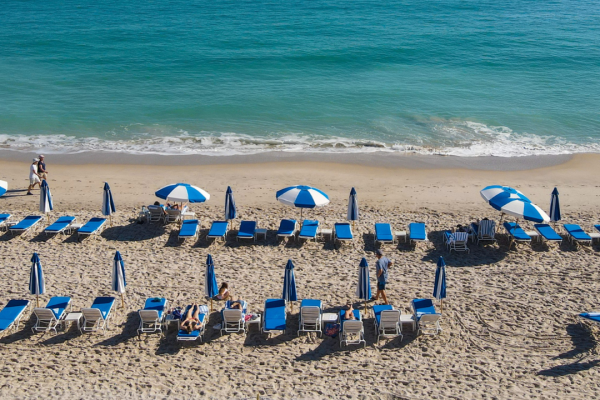 A beach scene with rows of blue and white lounge chairs and umbrellas, a few people are seen relaxing on the sand near the shoreline.