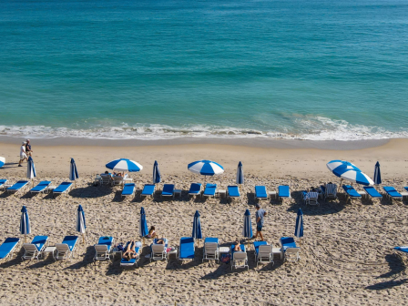 A beach scene with rows of blue and white lounge chairs and umbrellas, a few people are seen relaxing on the sand near the shoreline.