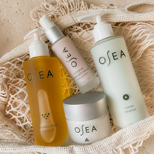 It shows a collection of Osea skincare products, including body oil, serum, cleanser, and protection cream, arranged in a net pouch on a textured surface.