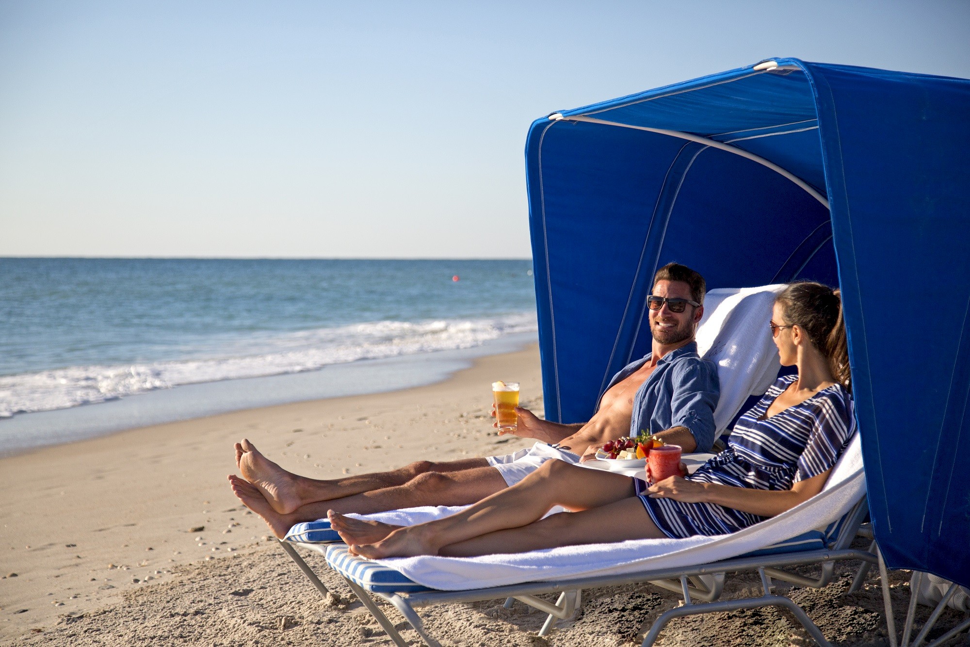 A couple relaxes on beach loungers under a blue canopy, drinks in hand, enjoying a sunny day by the ocean.