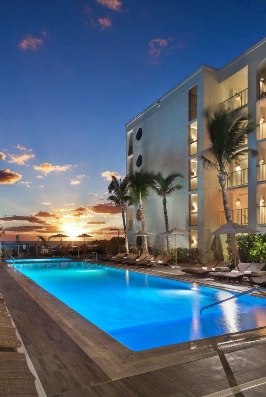 A luxurious hotel pool area during sunset with lounge chairs, umbrellas, palm trees, and a modern building in the background.