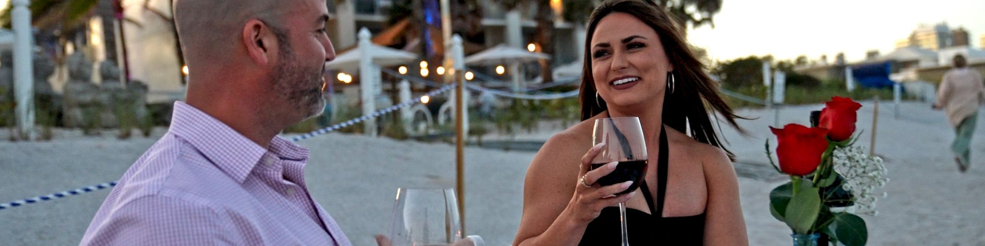 A man and a woman are enjoying a romantic dinner on a beach, holding wine glasses, with a table set with red roses, near a resort.