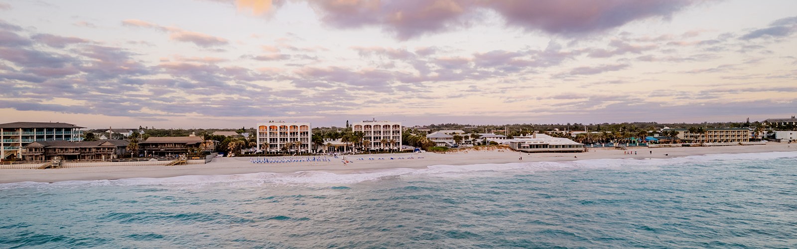 A scenic beach with gentle waves, lined with beachfront buildings under a sky with soft clouds during sunset or sunrise.